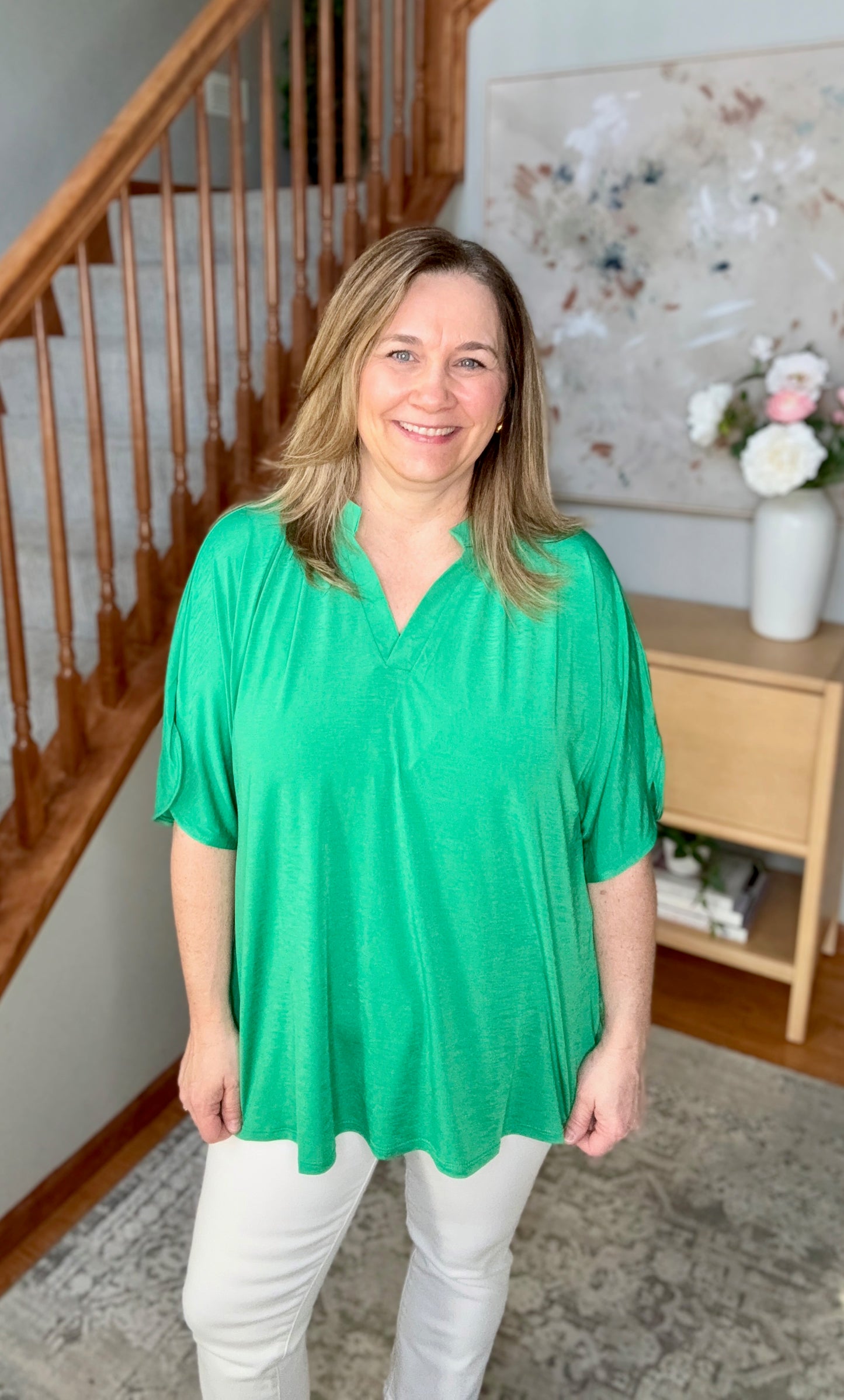 Woman wearing a green top and white pants standing in a home setting with a staircase and decorative items.