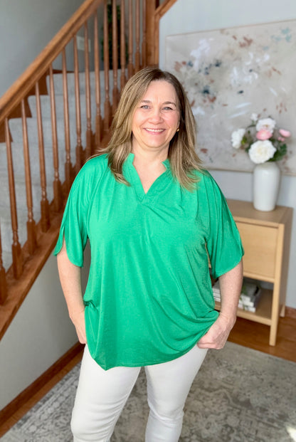 Woman in a green top and white pants standing in a home setting with a staircase and decor.
