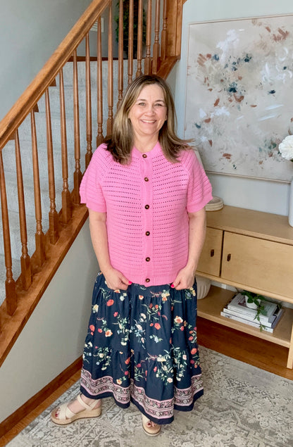Woman wearing a pink top and floral skirt standing in a home setting with a staircase and bookshelf.