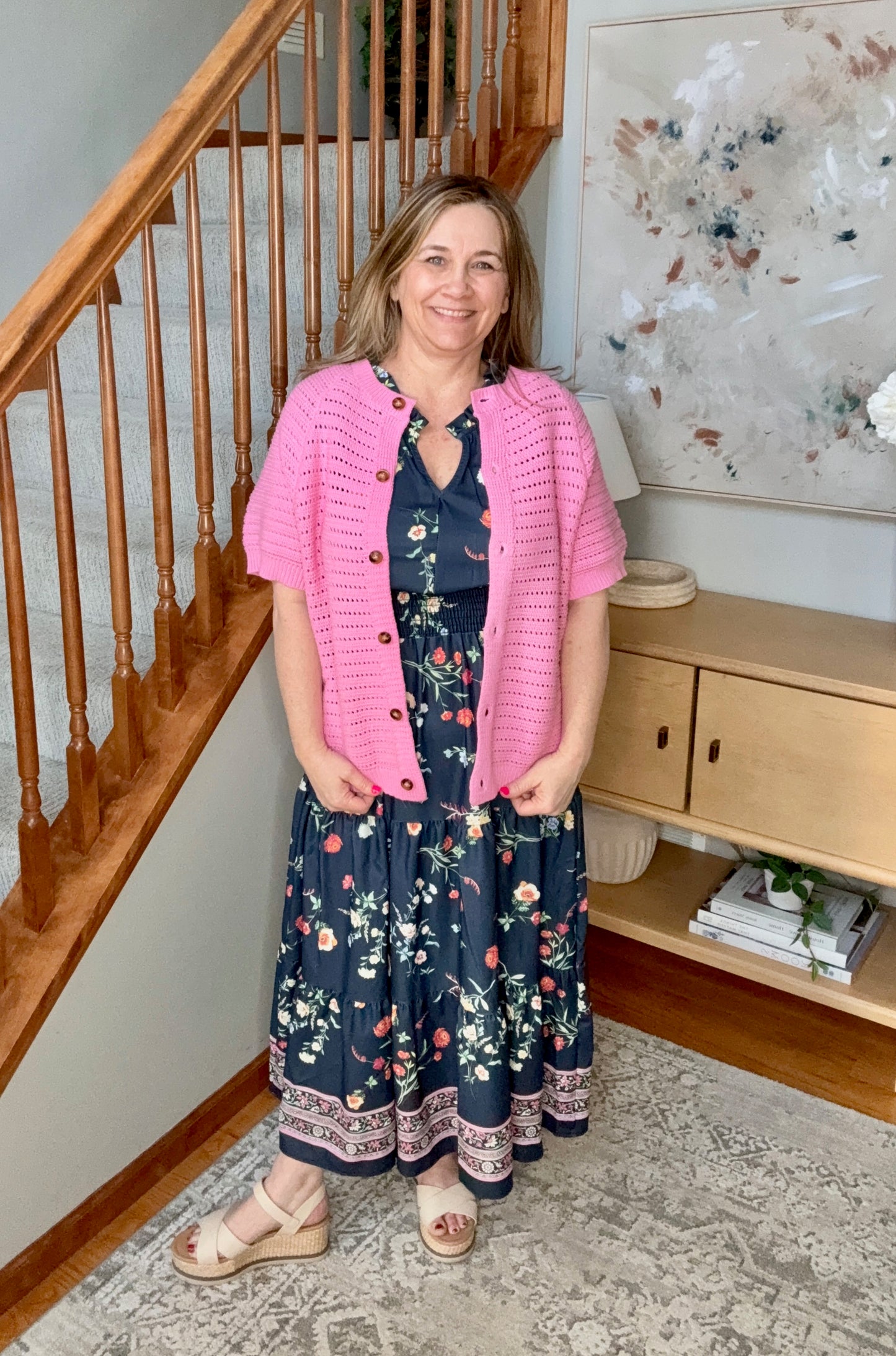 Woman wearing a pink cardigan over a floral dress standing in a home setting.