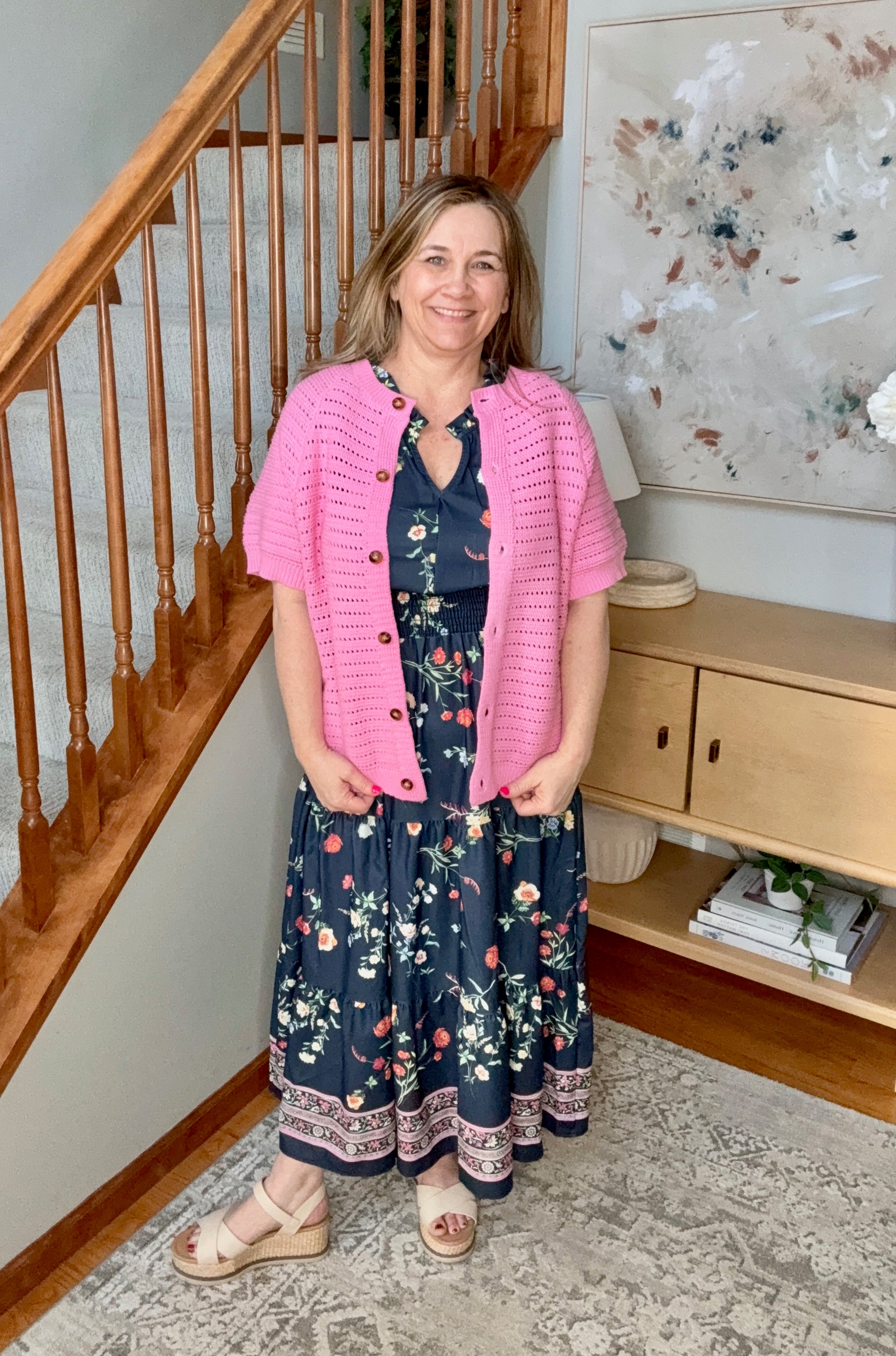 Woman wearing a pink cardigan over a floral dress standing in a home setting.