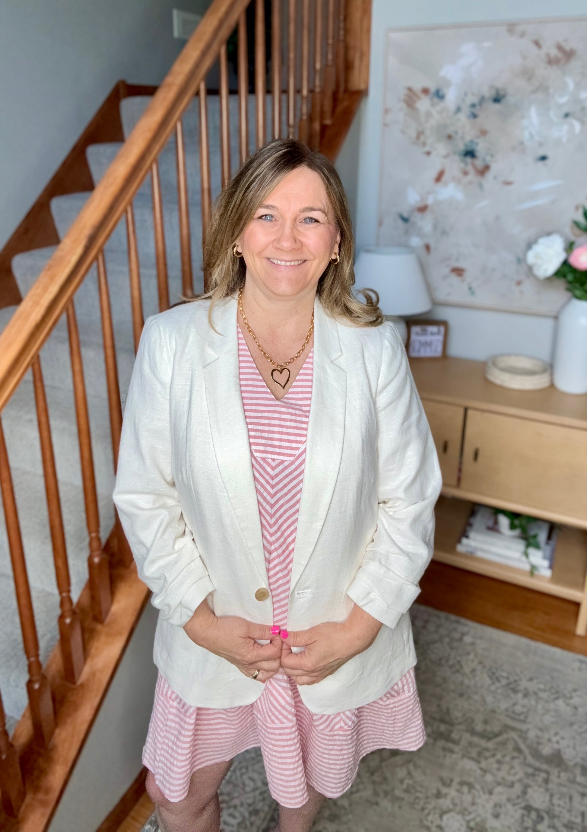 Woman in a white blazer and pink dress standing in a home setting with a staircase and decor.