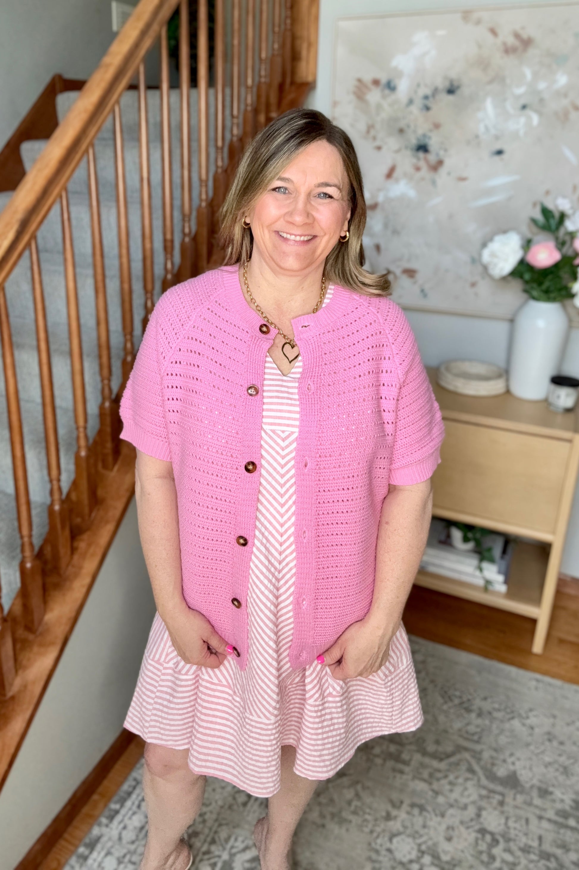 Woman wearing a pink cardigan over a dress in a home setting with a staircase and decor.