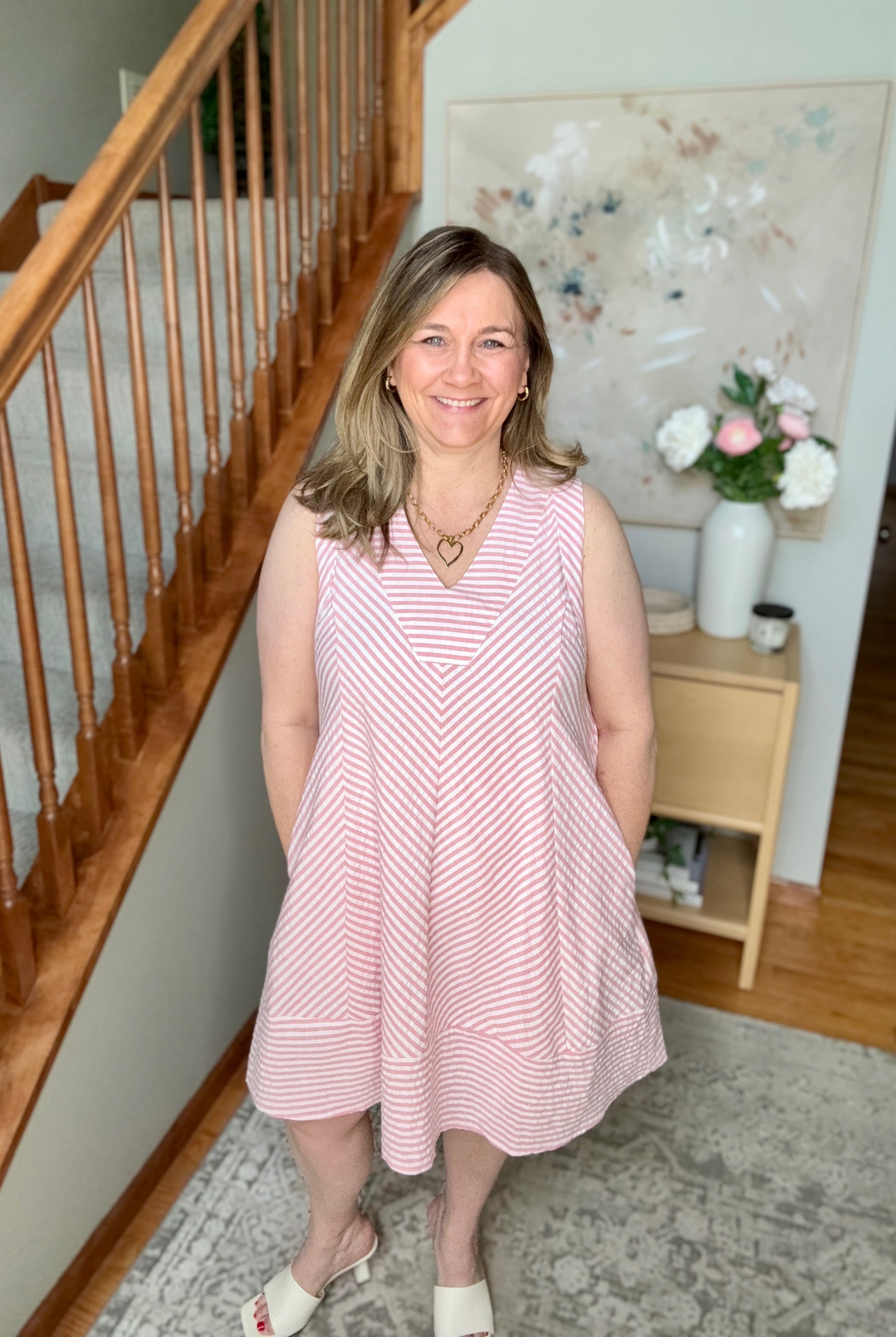 Woman in a pink dress standing in a home interior with wooden staircase and floral decor.