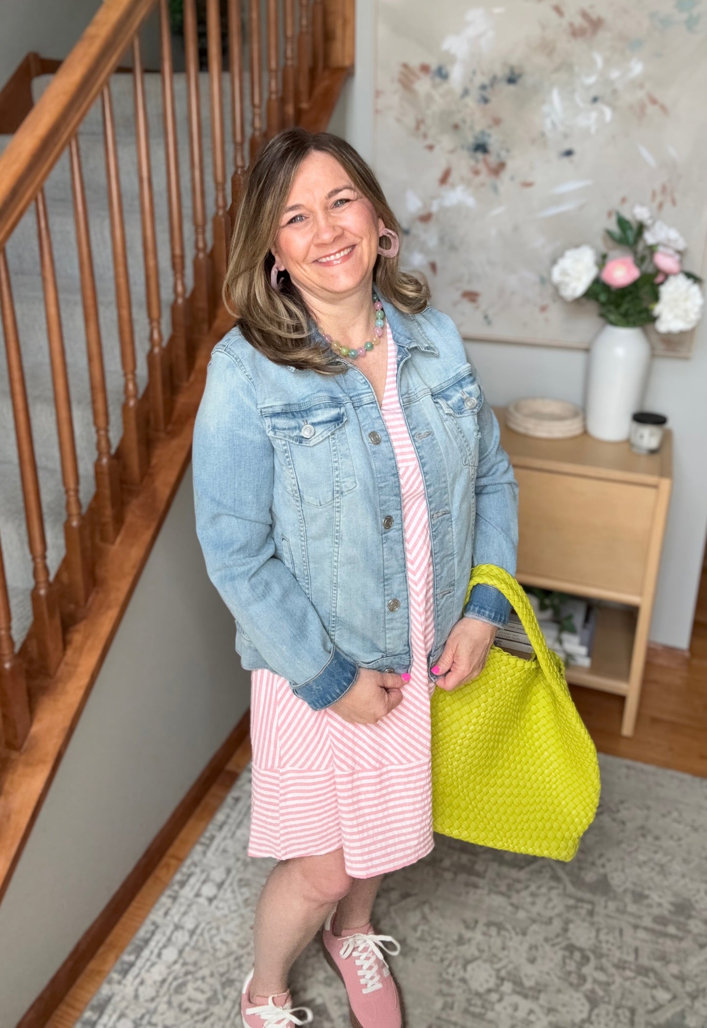 Woman in a denim jacket and pink dress holding a yellow bag in a home setting.