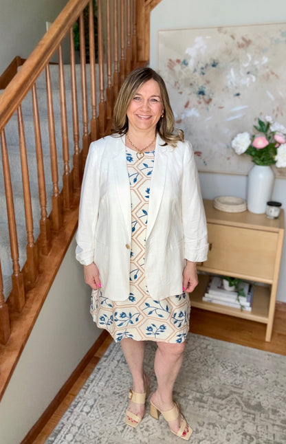 Woman in a white jacket and patterned dress standing in a home setting with wooden staircase and decor.