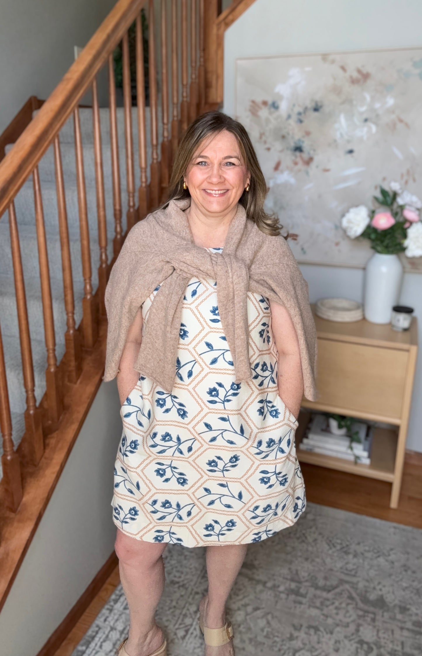 Woman standing in a home setting with wooden staircase and floral decorations.