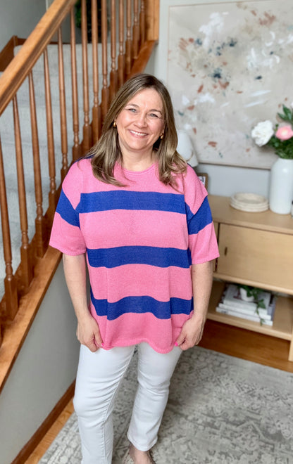 Woman wearing a pink and blue striped shirt standing in a home setting with a staircase and decor.
