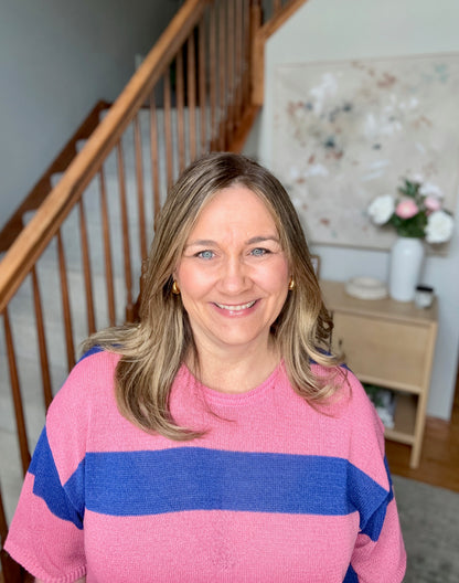 Woman wearing a pink and blue sweater standing in a home setting with a staircase and decor.