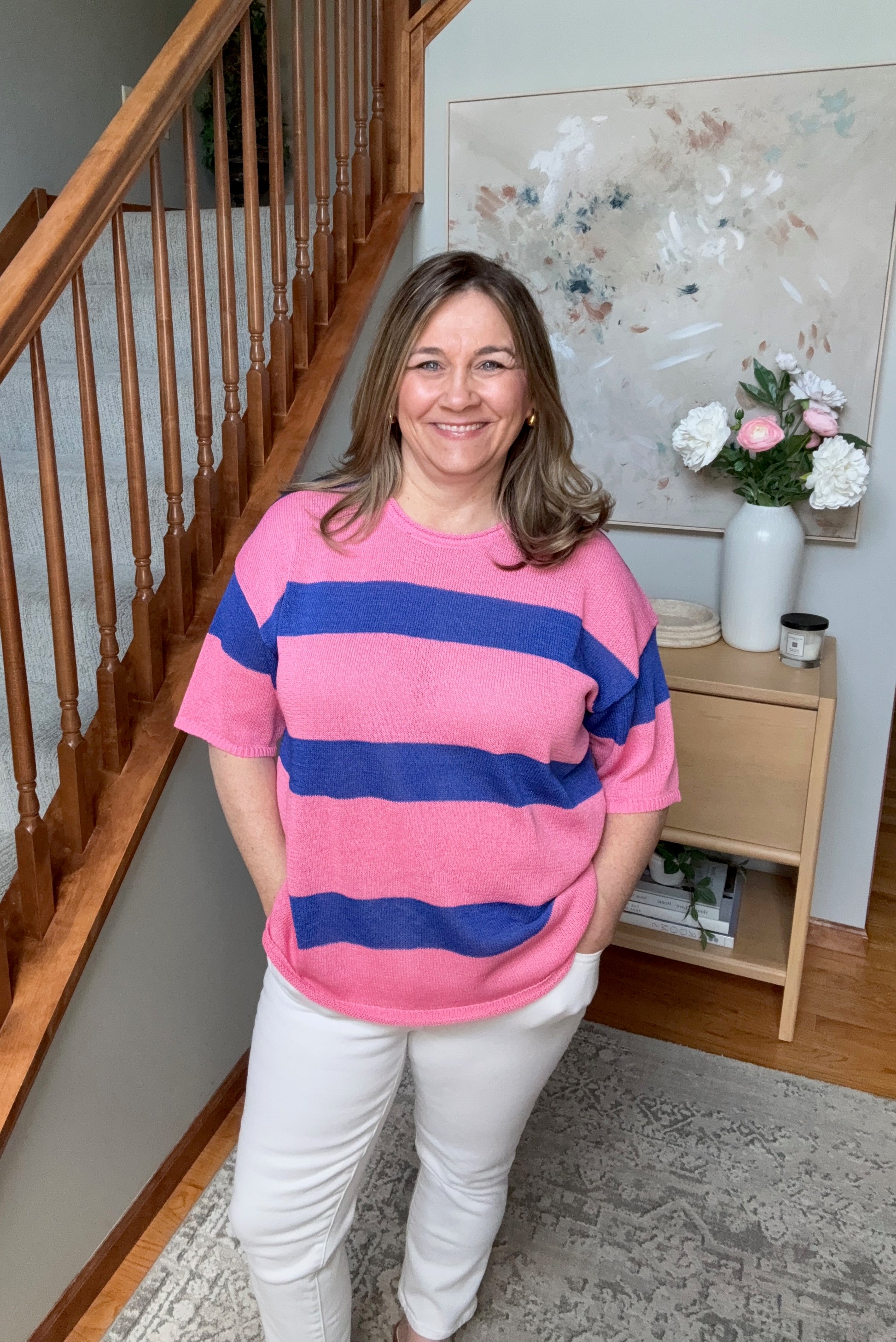 Woman wearing a pink and blue striped sweater standing in a home setting with a staircase and artwork in the background.
