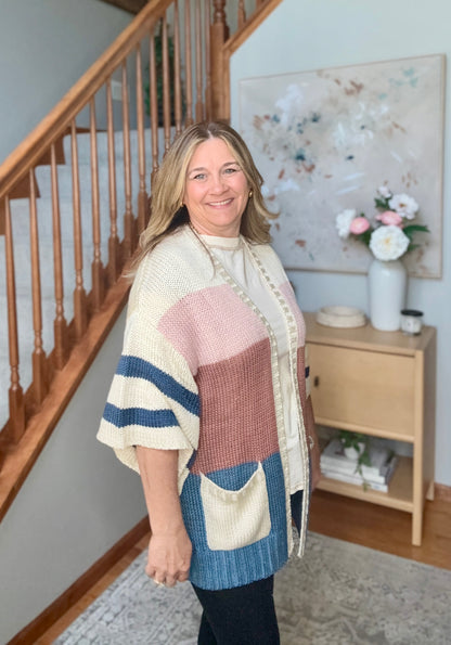 A woman wearing a chunky knit color-block poncho cardigan with cream, dusty pink, navy blue, and light blue panels, standing in a home entryway next to a wooden staircase.