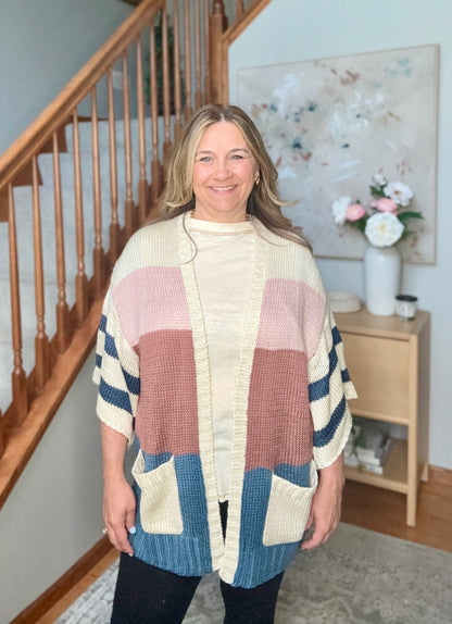 A woman wearing a chunky knit color-block poncho cardigan with cream, dusty pink, terracotta, and blue panels, standing in a home interior with a staircase and sideboard in the background.
