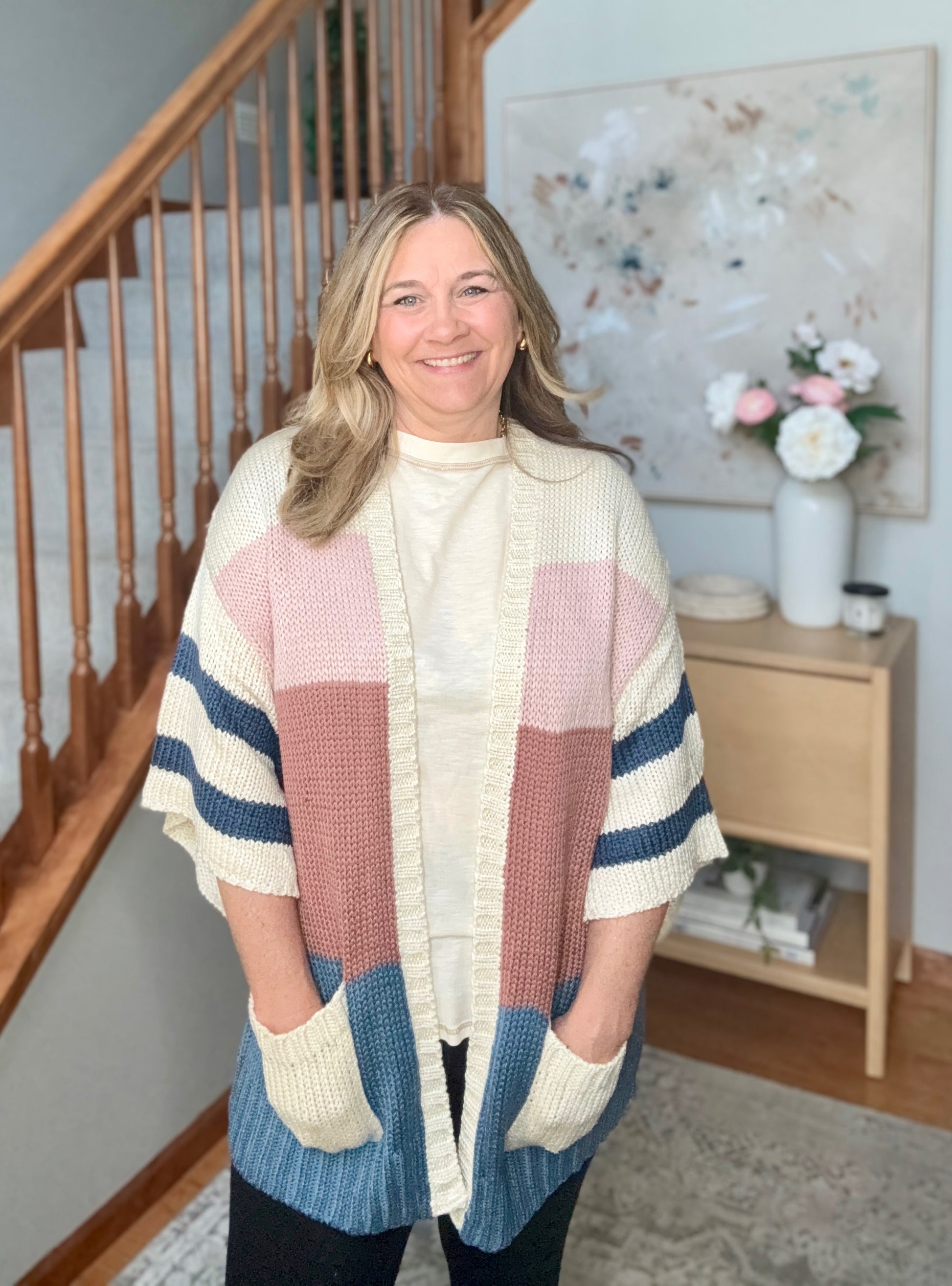 A woman wearing a chunky knit cardigan with large color-blocked panels in cream, dusty pink, terracotta, and blue, standing in a home setting with a staircase and side table in the background.