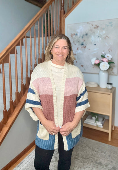 A woman stands indoors wearing a chunky knit color-block poncho sweater with cream, dusty pink, mauve, and blue panels, photographed in a home setting with a staircase and side table in the background.