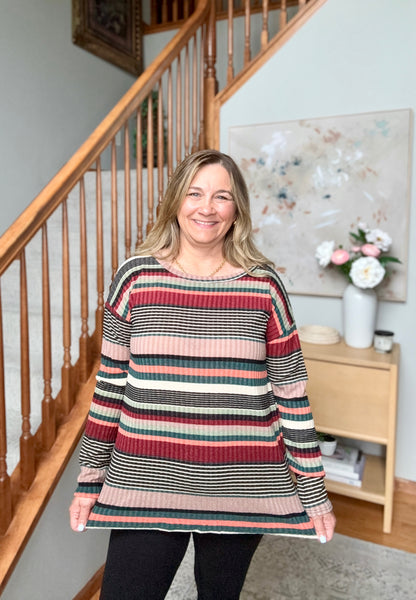 A woman wearing a long-sleeve, crewneck ribbed sweater with horizontal stripes in red, pink, green, black, and white, standing in a home interior in front of a wooden staircase.