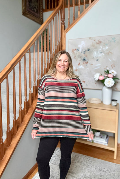 A woman stands in a home interior in front of a wooden staircase, wearing a long-sleeve knit sweater with horizontal stripes in red, beige, black, and teal, paired with black leggings.