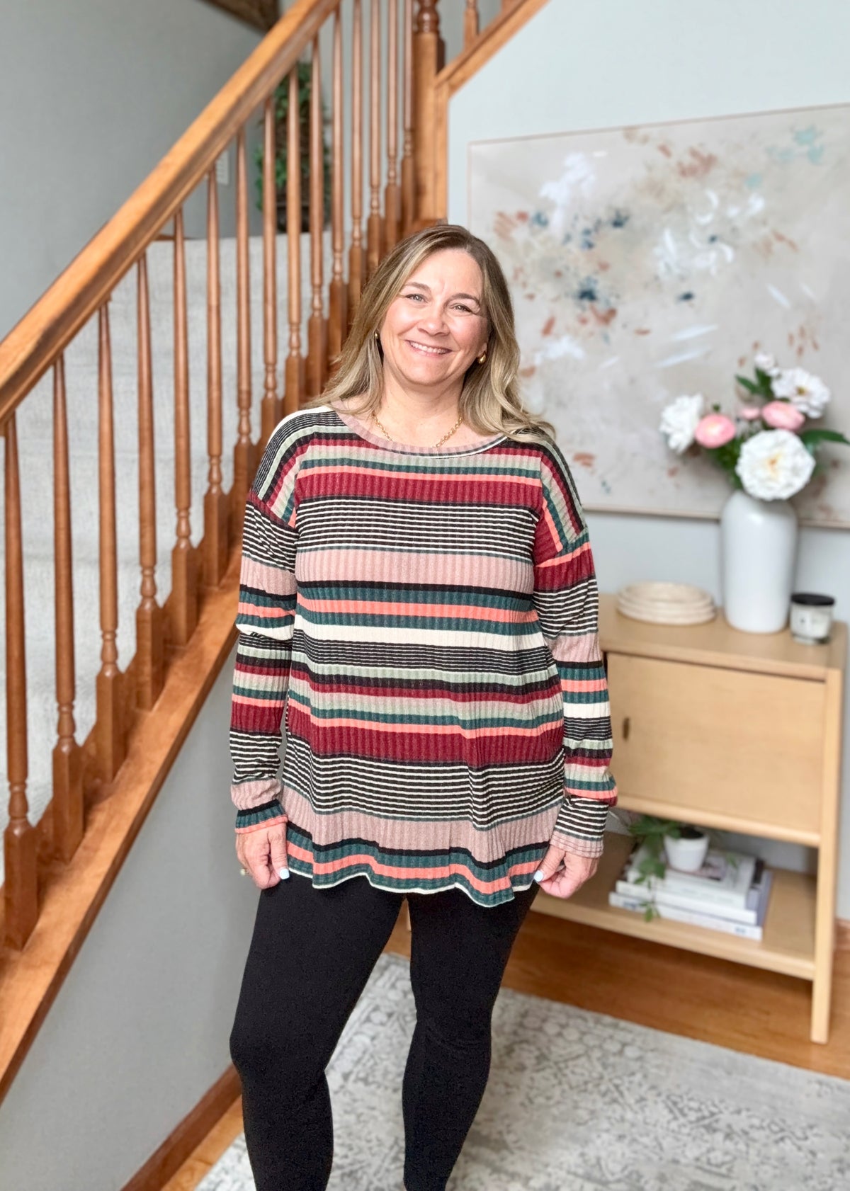 A woman stands in a home interior wearing a long-sleeve knit sweater with horizontal stripes in red, pink, black, and beige, paired with black leggings.