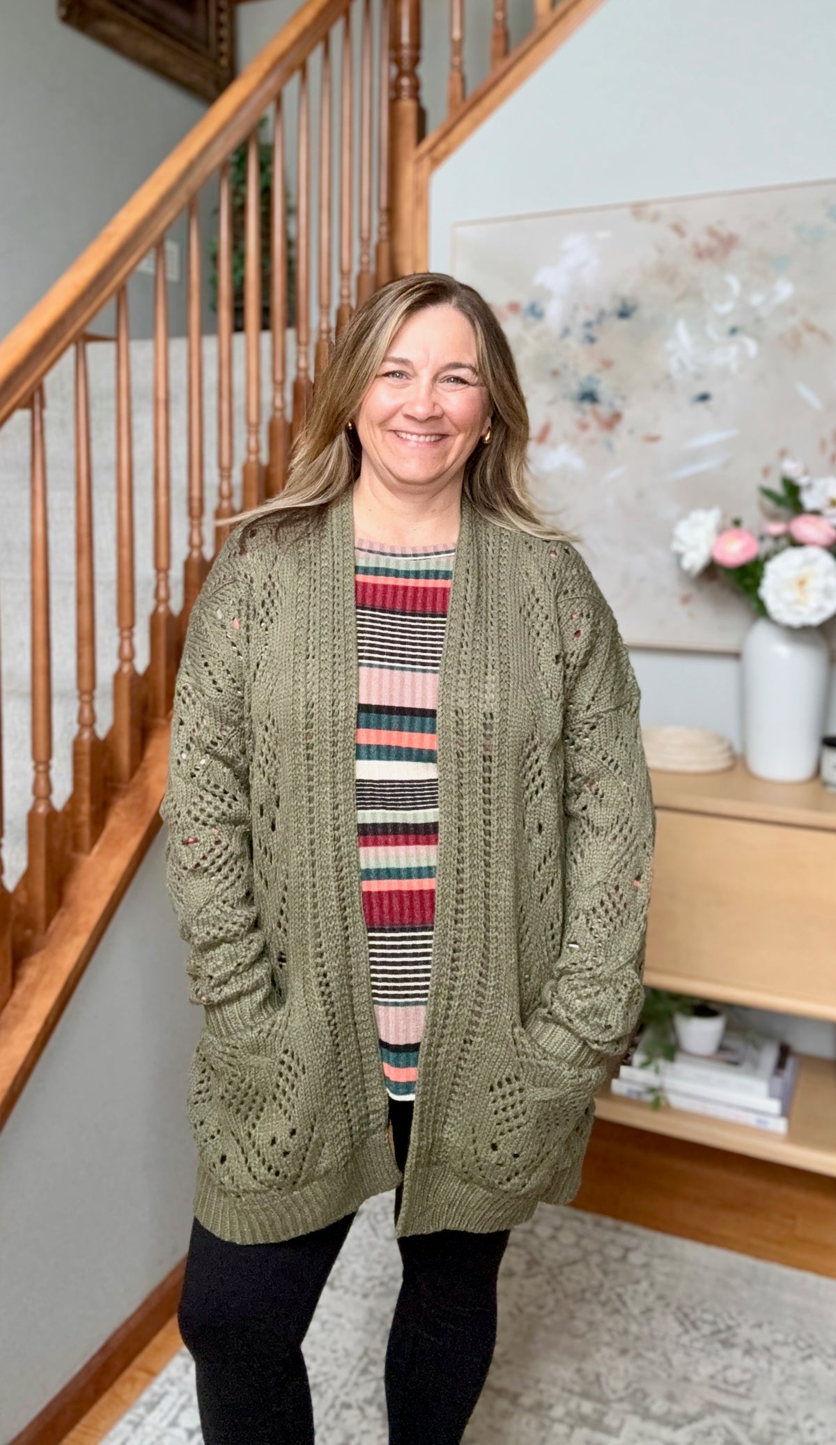 A smiling woman stands indoors wearing a loose, long-sleeve olive green cable-knit cardigan over a striped sweater. The setting is a home interior with a wooden staircase, light walls, and a side table in the background.