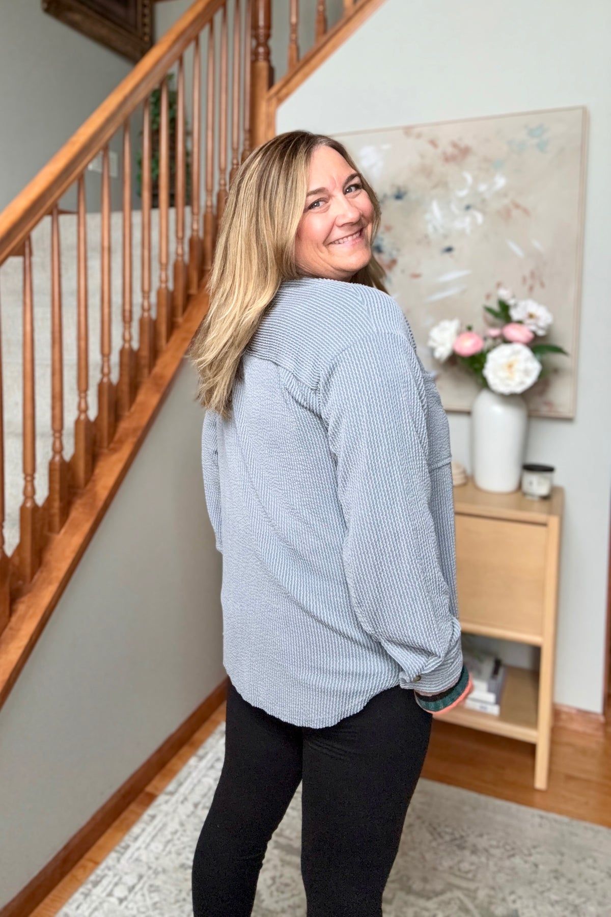 A woman with blonde hair wearing a light blue-gray ribbed long-sleeve top and black pants, standing in a home interior near a wooden staircase and a side table with flowers.