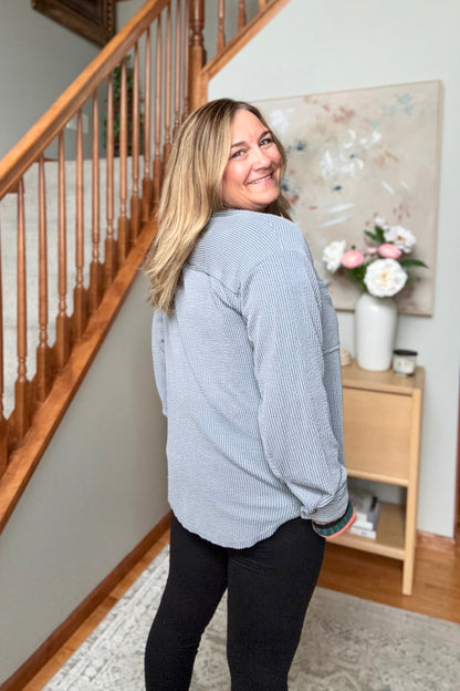A woman with blonde hair wearing a light blue-gray ribbed long-sleeve top and black pants, standing in a home interior near a wooden staircase and a side table with flowers.