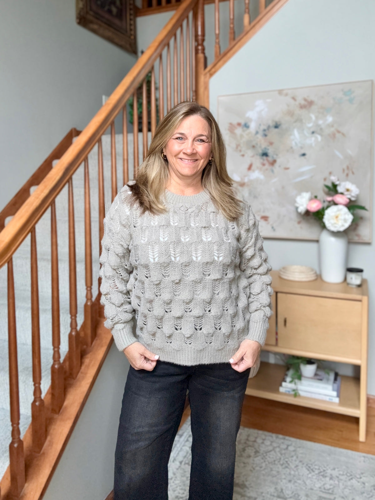 A woman stands indoors in front of a wooden staircase, wearing a light gray cable-knit sweater and dark jeans.