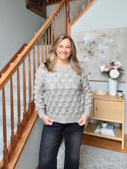 A woman stands indoors in front of a wooden staircase, wearing a light gray cable-knit sweater and dark jeans.