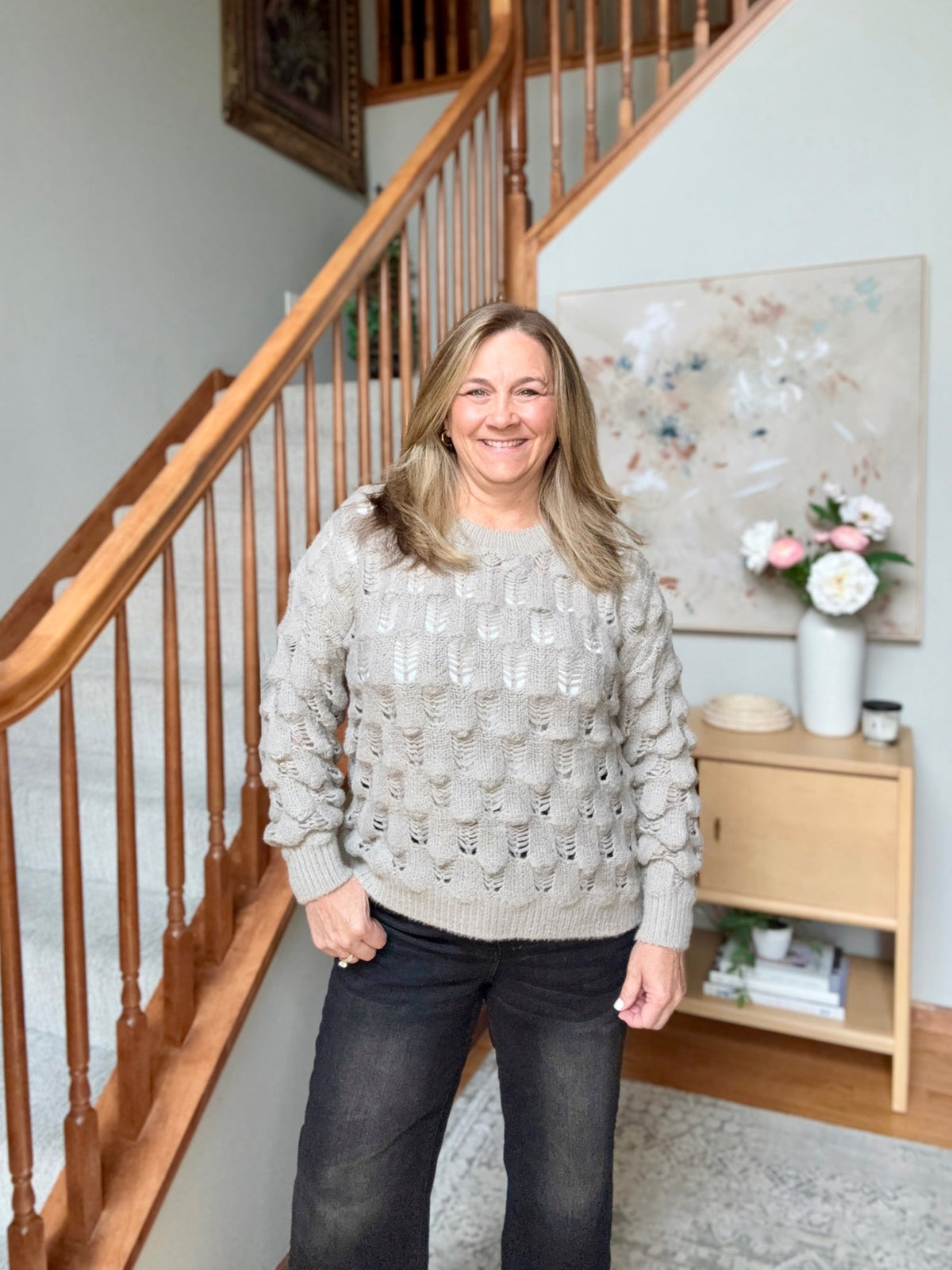 A smiling woman with blonde hair stands on a staircase landing wearing a light beige textured cable-knit sweater and dark gray jeans. A wooden handrail is on the left, and a small wooden side table with a vase of flowers is on the right.