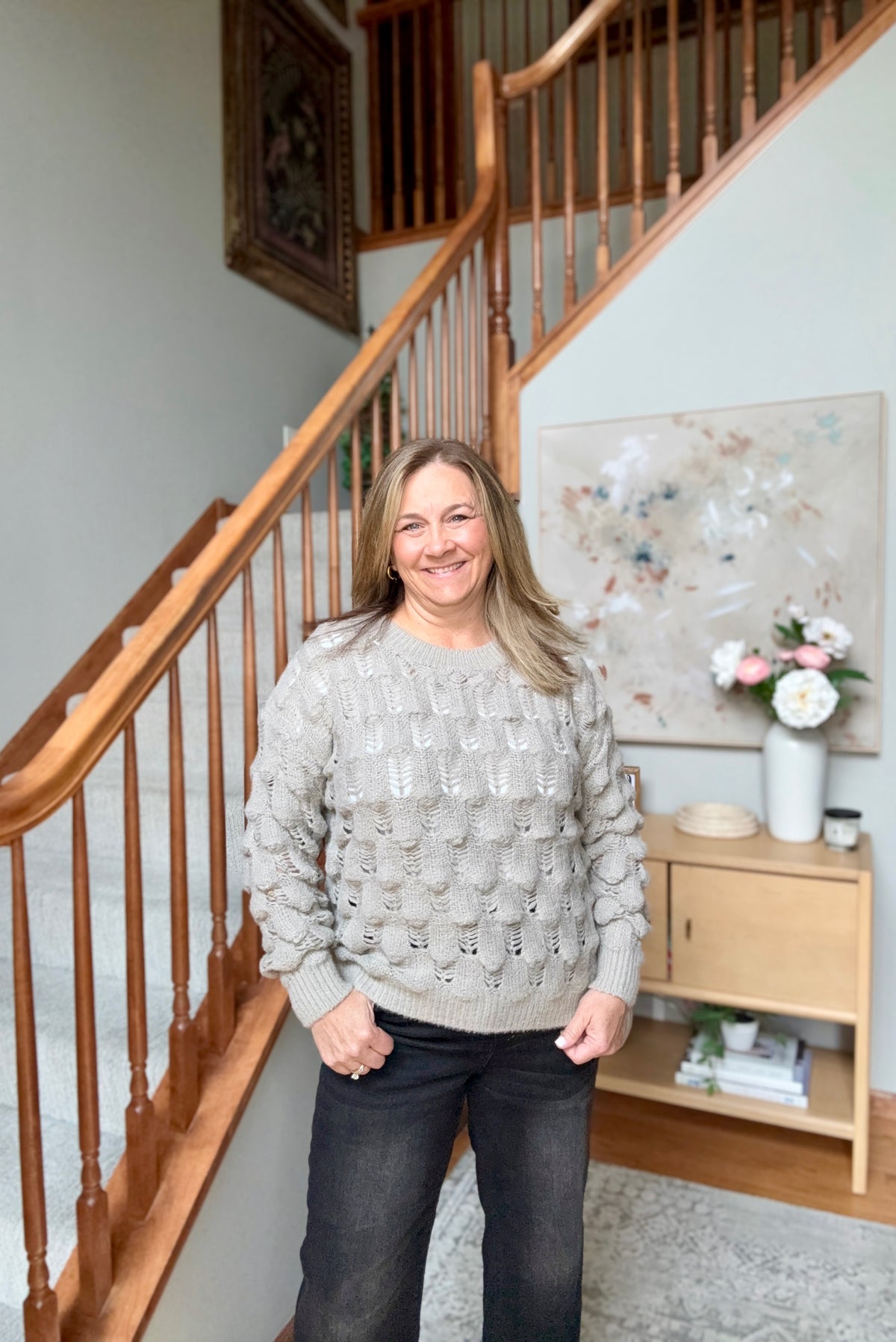 A smiling woman stands indoors in front of a wooden staircase, wearing a light gray textured knit sweater and dark jeans. The setting includes a small wooden side table with flowers and framed wall art.
