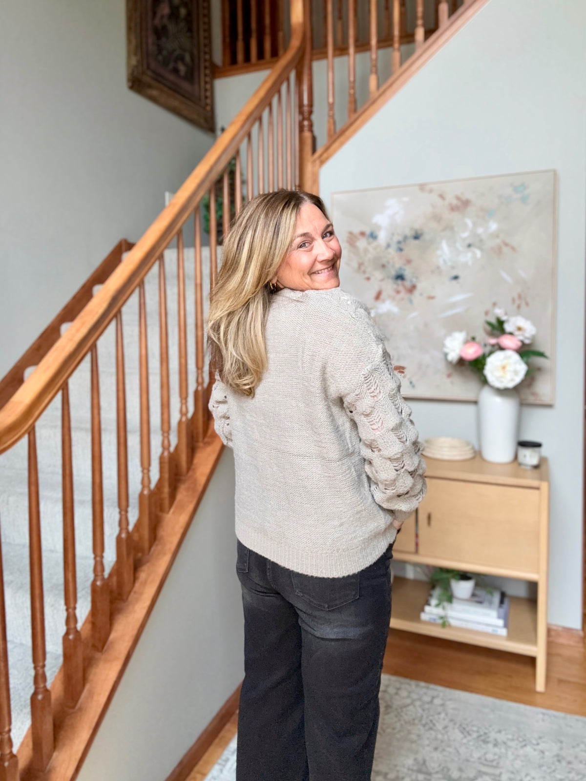 A woman with blonde hair stands in a home interior next to a wooden staircase, wearing a light beige textured knit sweater with decorative cable-knit sleeves and dark gray pants.