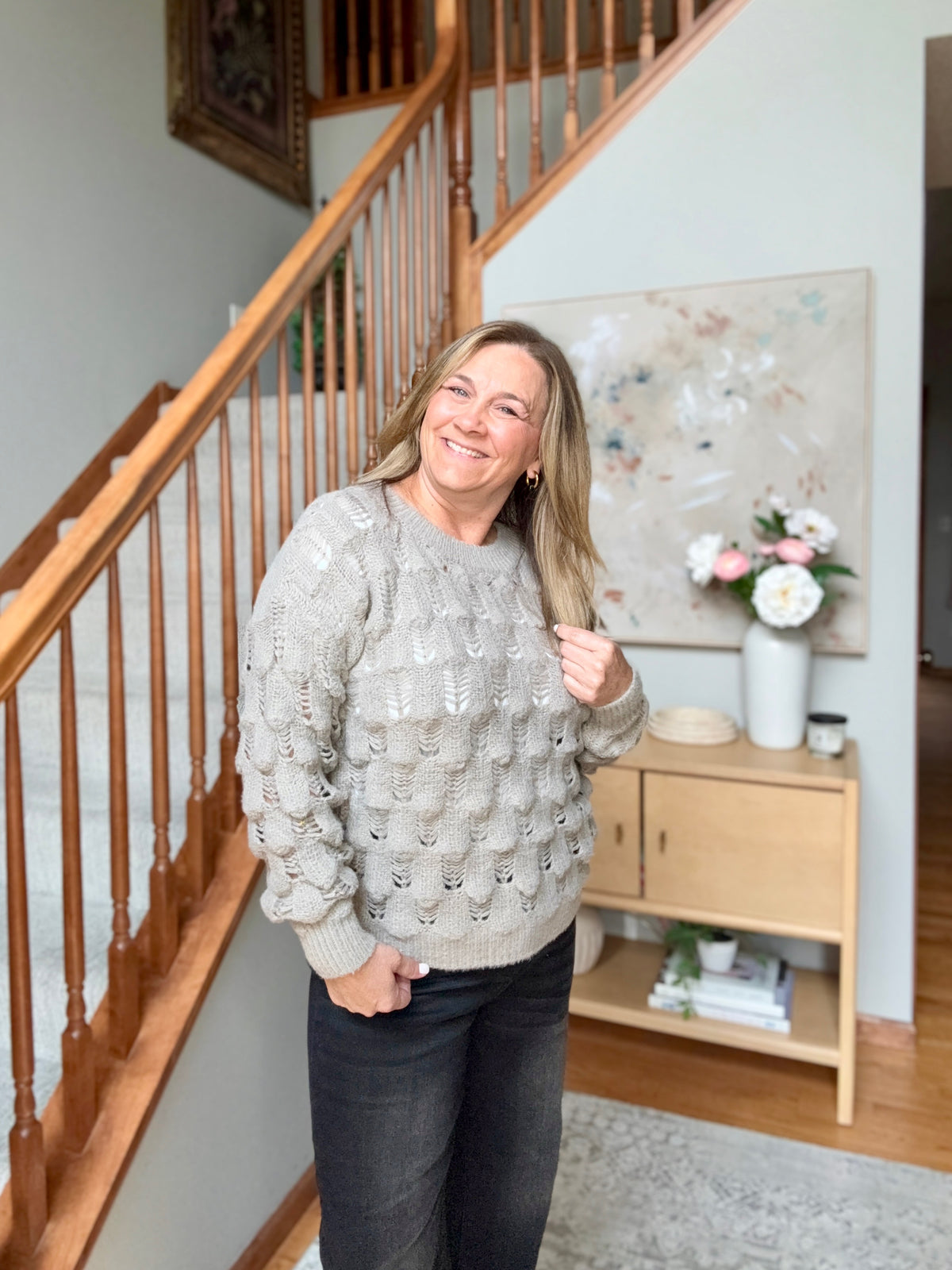 A woman smiling while wearing a light gray cable-knit sweater and dark jeans, standing in a home entryway next to a wooden staircase and a small wooden sideboard with a vase of flowers.