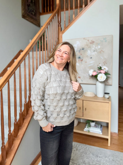 A woman smiling while wearing a light gray cable-knit sweater and dark jeans, standing in a home entryway next to a wooden staircase and a small wooden sideboard with a vase of flowers.