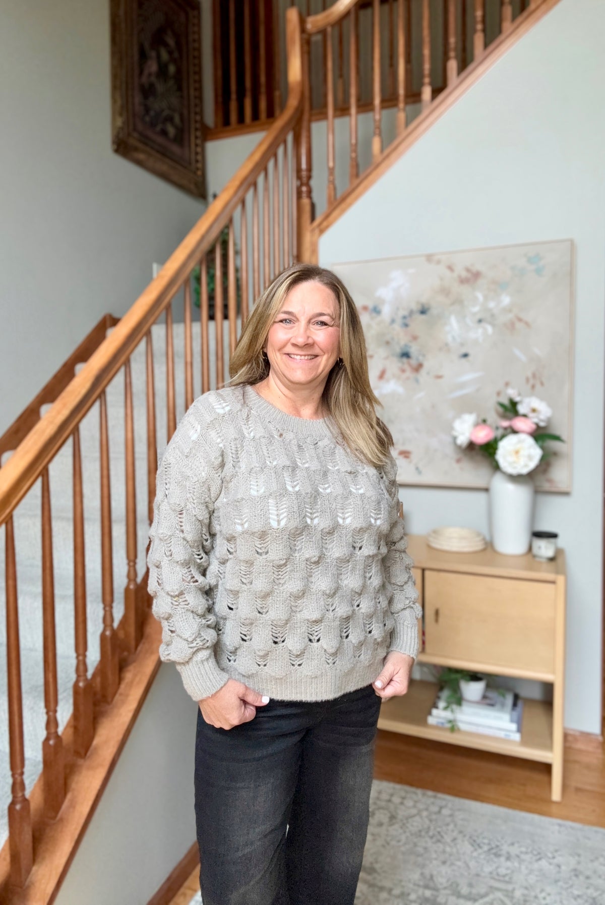 A smiling woman stands indoors in front of a wooden staircase, wearing a light gray textured knit sweater and dark jeans. A small wooden cabinet with flowers is visible in the background.