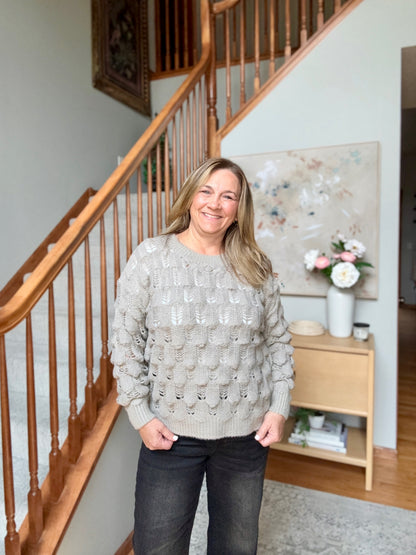 A smiling woman stands indoors in front of a wooden staircase, wearing a light taupe-gray textured knit sweater and dark jeans.