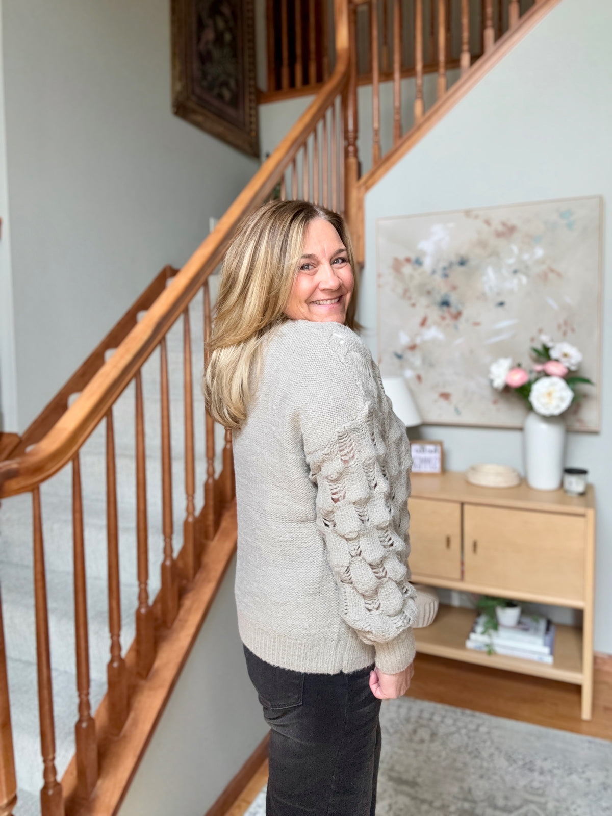 A woman with blonde hair stands on a staircase landing, looking back over her shoulder while wearing a light beige textured knit sweater with a raised bubble pattern on the sleeves and dark pants.