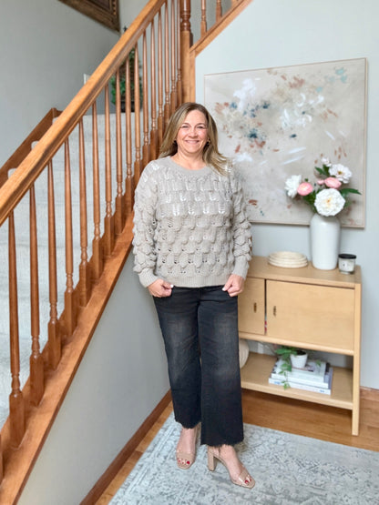 A woman stands in a home interior next to a wooden staircase, wearing a textured light beige knit sweater and dark cropped jeans.