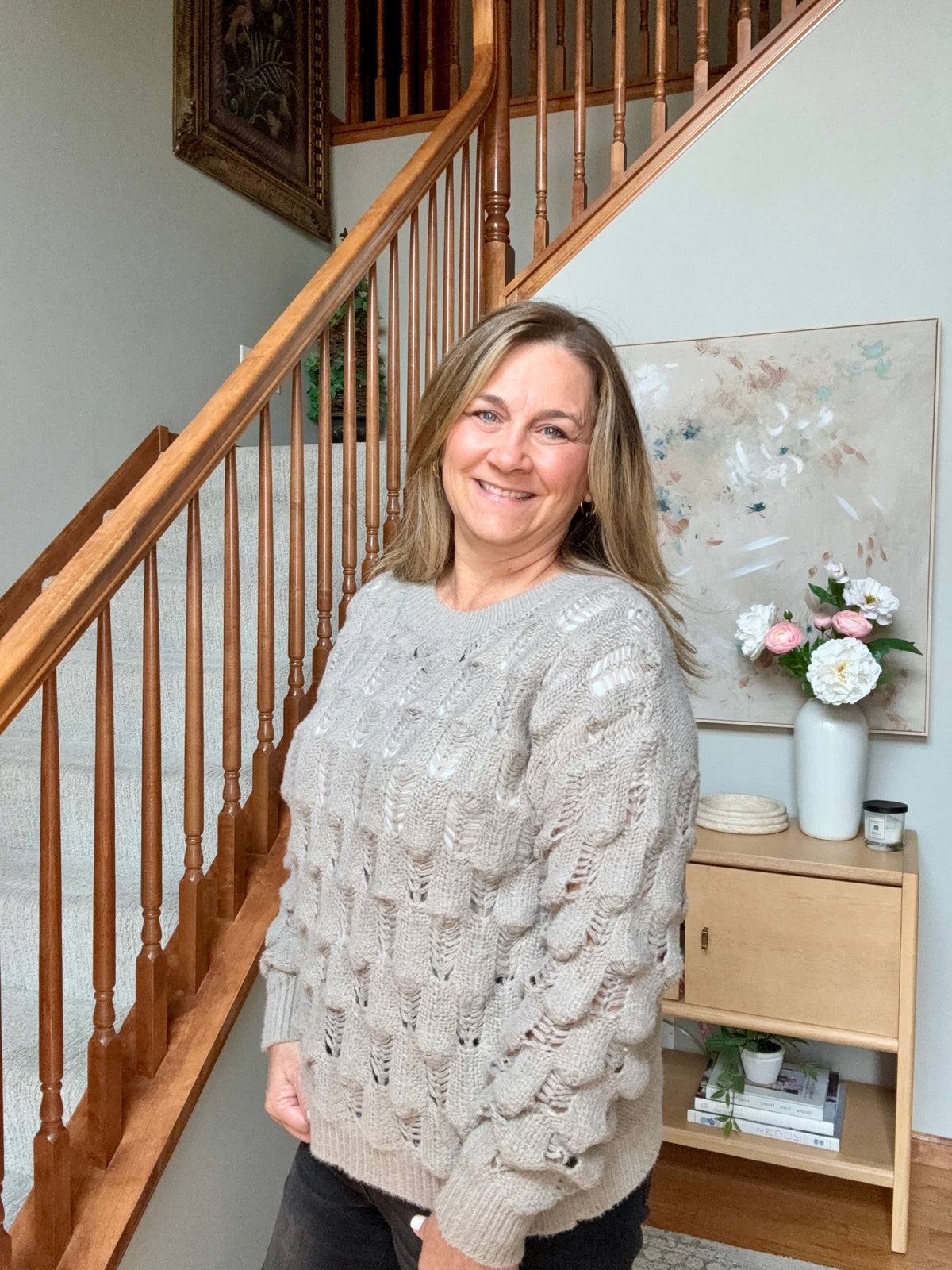 A woman smiling while wearing a chunky, textured cable-knit sweater in a light taupe-beige color, standing in a home interior next to a wooden staircase.