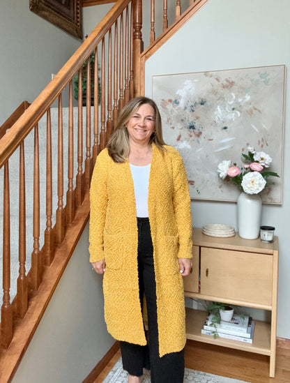 A woman stands in a home interior wearing a textured mustard yellow open-front cardigan over a white top and black pants. A wooden staircase, sideboard with flowers, and framed wall art are visible in the background.