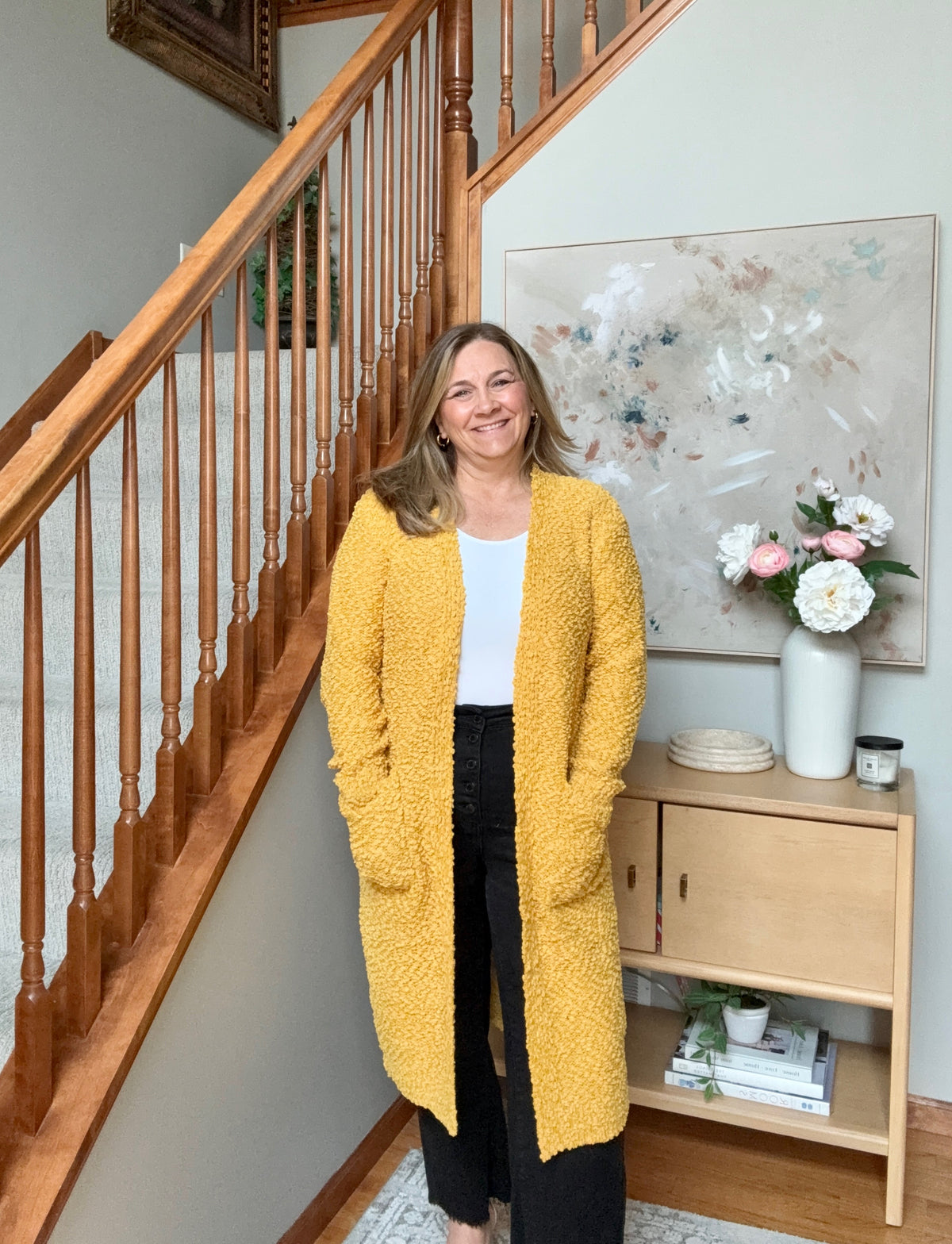 A woman stands in a home interior next to a wooden staircase, wearing a textured mustard yellow open-front cardigan over a white top and black pants. A light wood console table with flowers and a large abstract painting is visible in the background.