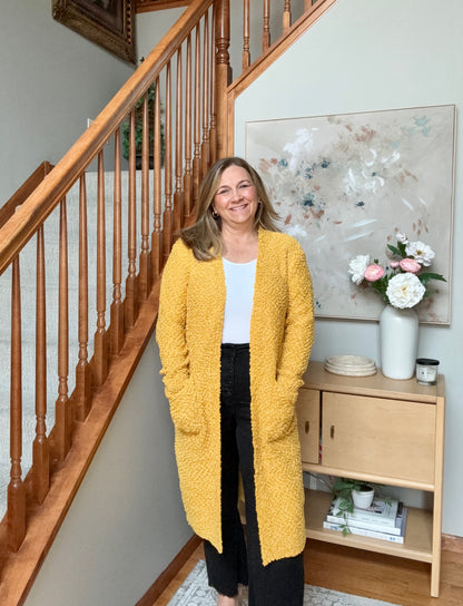 A woman stands in a home interior next to a wooden staircase, wearing a textured mustard yellow open-front cardigan over a white top and black pants. A light wood console table with flowers and a large abstract painting is visible in the background.