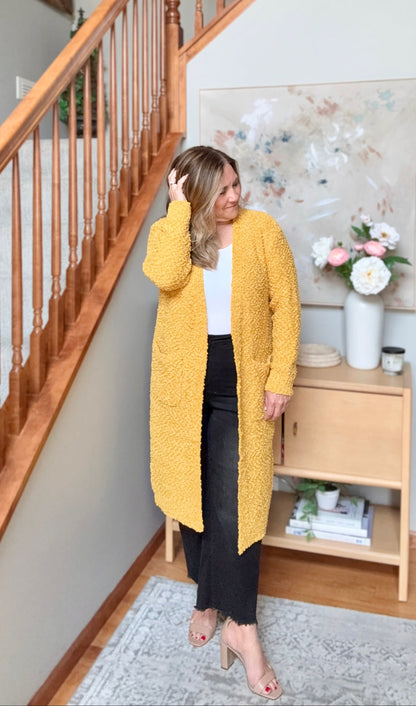 A woman wearing a textured mustard yellow cardigan over a white top and black pants, standing in a home hallway next to a wooden staircase.