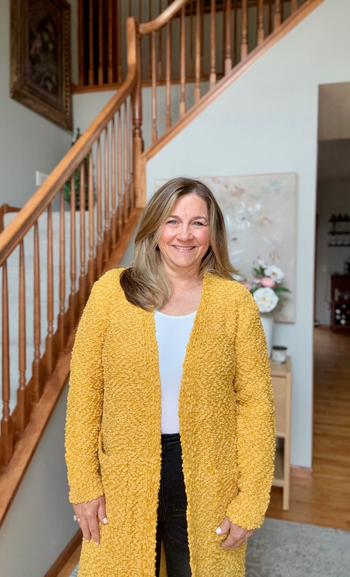 A smiling woman with blonde hair stands indoors in front of a wooden staircase, wearing a textured mustard yellow open-front cardigan over a white top and black pants.