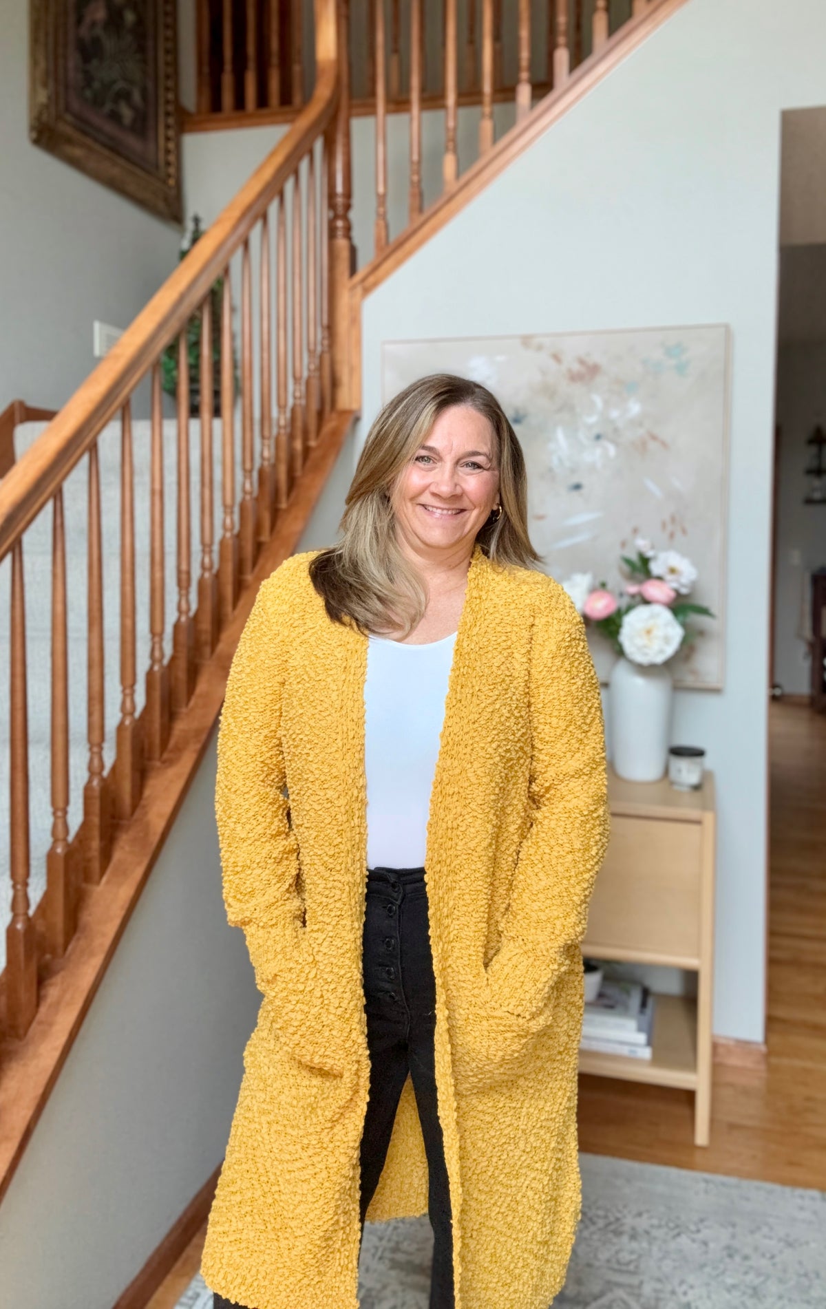 A woman stands in a home interior wearing a long, open-front textured cardigan in mustard yellow over a white top and black pants. The background features a wooden staircase, a light wood side table with flowers, and a neutral wall.