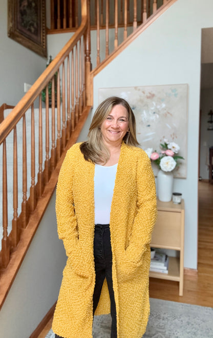 A woman stands in a home interior wearing a long, open-front textured cardigan in mustard yellow over a white top and black pants. The background features a wooden staircase, a light wood side table with flowers, and a neutral wall.