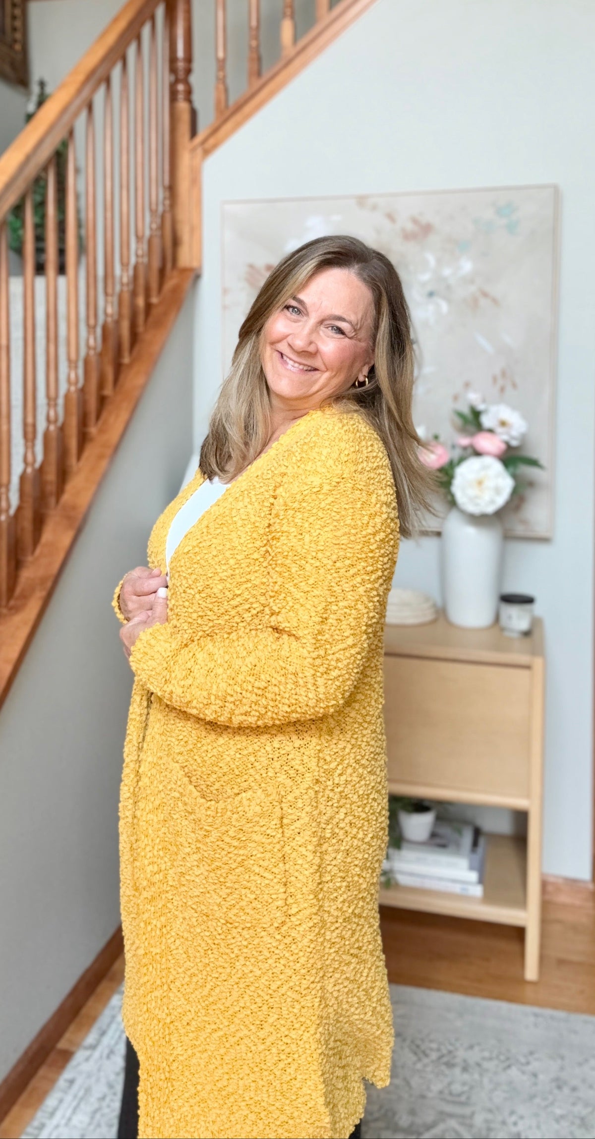A smiling woman stands in a home interior near a wooden staircase, wearing a bright yellow textured cardigan over a white top. A small side table with flowers and decor is visible in the background.