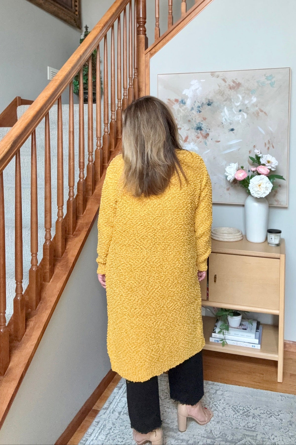 A woman viewed from behind wearing a textured mustard yellow long-sleeve cardigan, standing in a home entryway next to a wooden staircase and a small wooden side table.