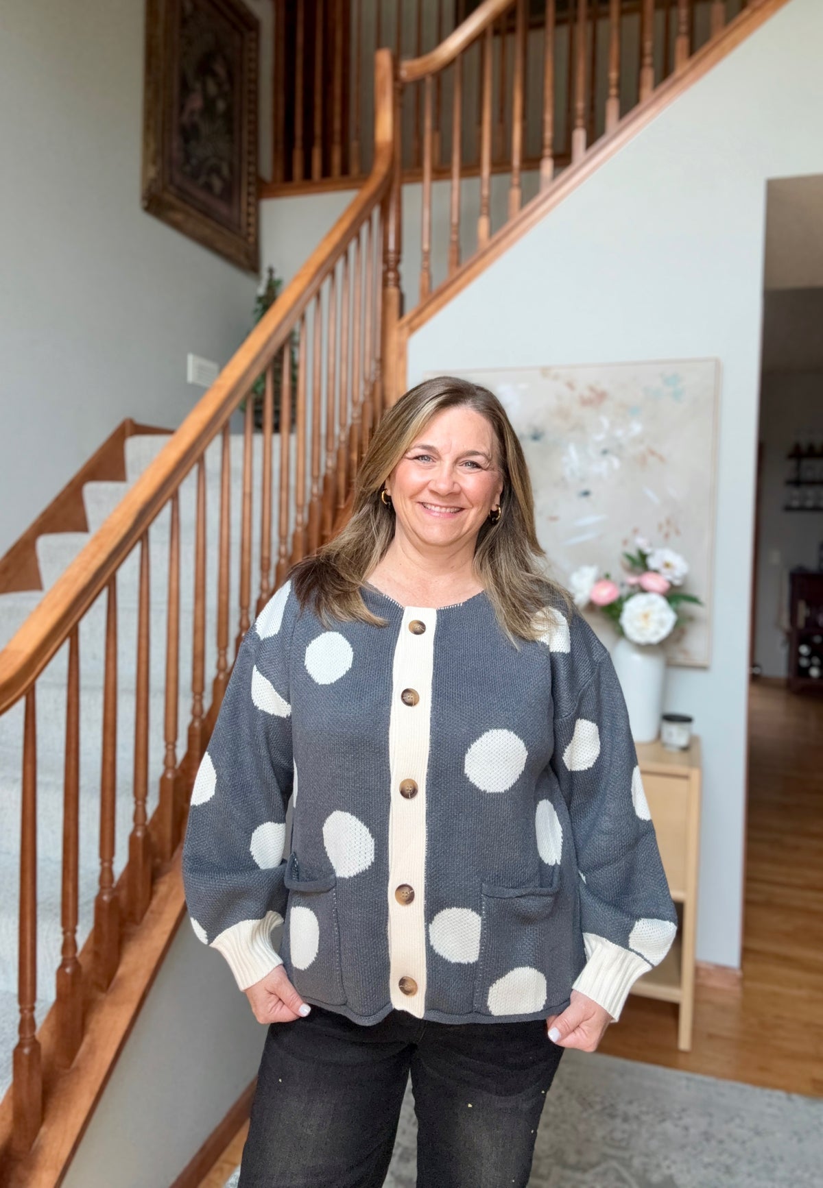 A woman stands in front of a wooden staircase wearing a gray cardigan with large white polka dots and a cream button placket. She is also wearing dark jeans.