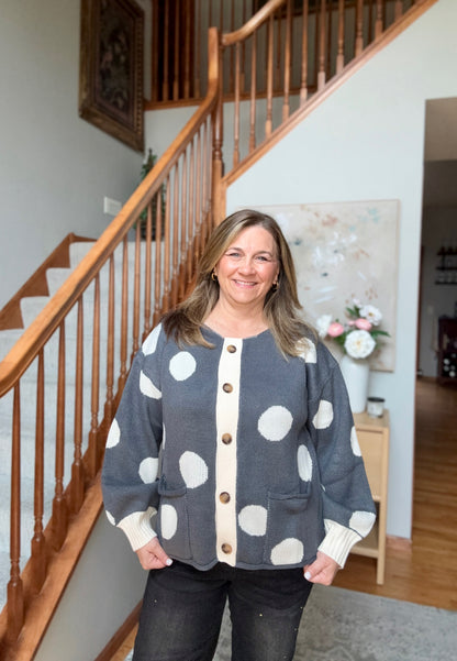 A woman stands in front of a wooden staircase wearing a gray cardigan with large white polka dots and a cream button placket. She is also wearing dark jeans.