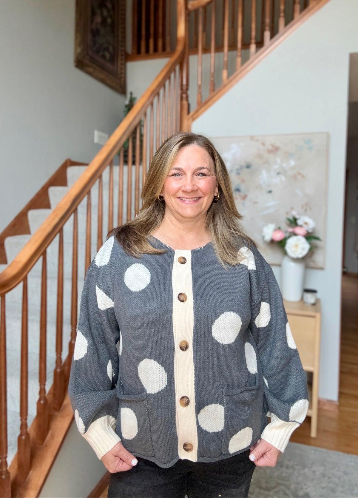 A woman smiling in a home interior, wearing a gray knit cardigan with large white polka dots, a cream center placket, and brown buttons. The background shows a wooden staircase and a home hallway.