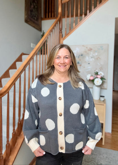 A woman smiling in a home interior, wearing a gray knit cardigan with large white polka dots, a cream center placket, and brown buttons. The background shows a wooden staircase and a home hallway.