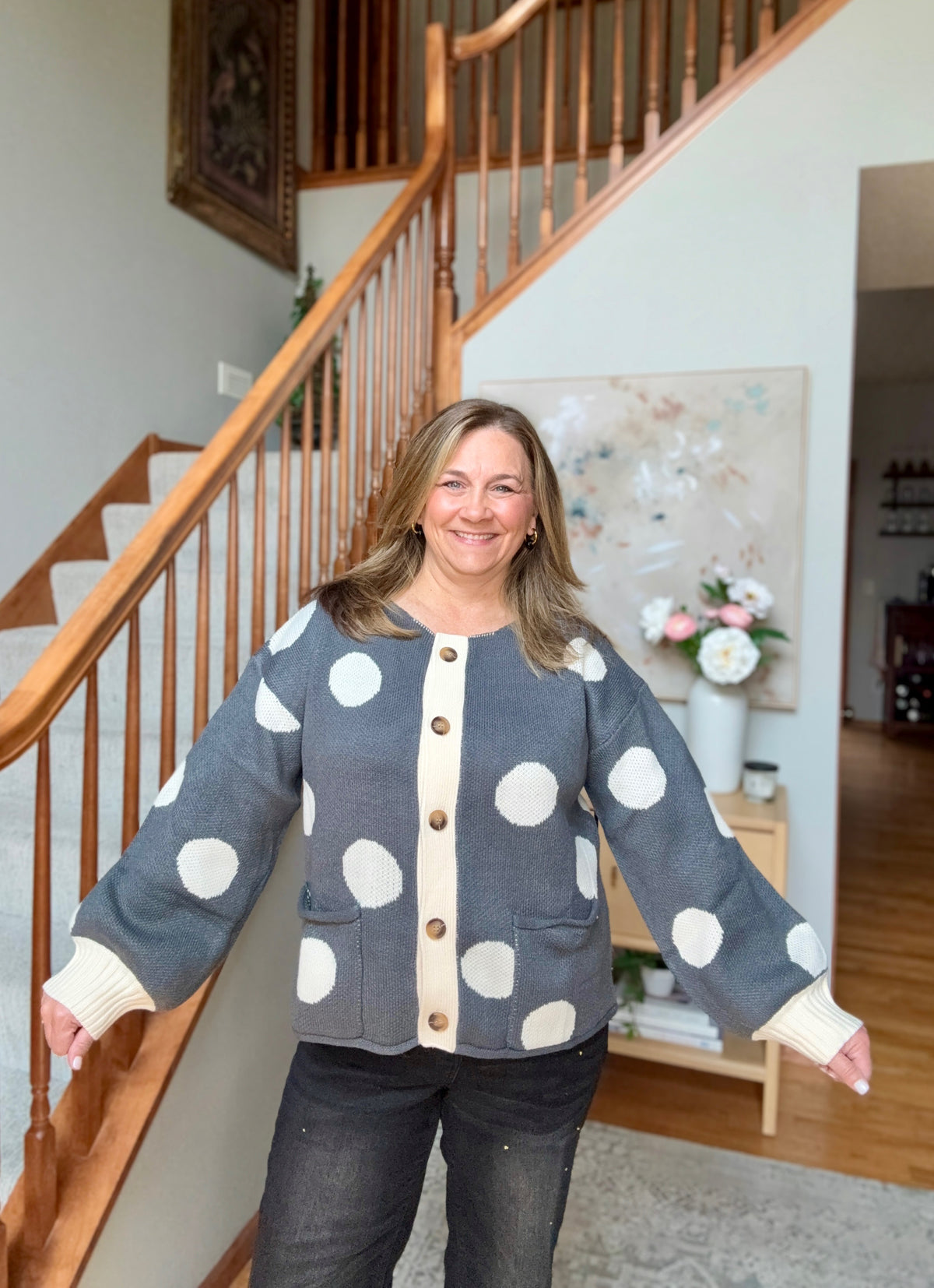 A woman stands in a home interior in front of a wooden staircase, wearing a gray cardigan with large white polka dots and a cream button placket. The cardigan has ribbed cuffs and a front pocket.